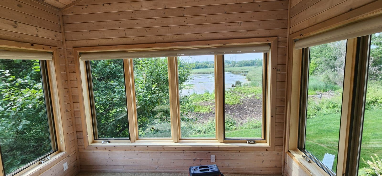 Knotty-pine sunroom corner with new wood casement windows looking out at a pond